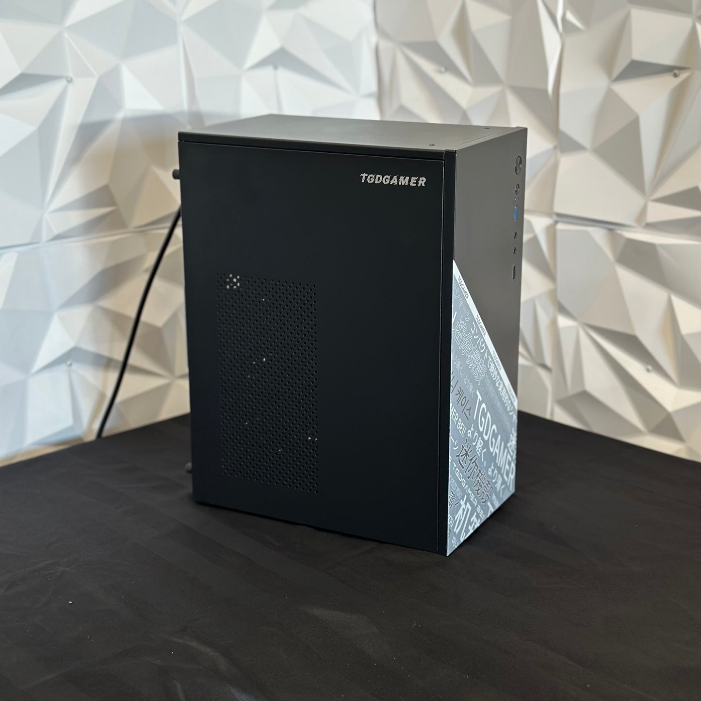 Black computer case with 'TGDGAMER' branding on a black surface and geometric-patterned wall background