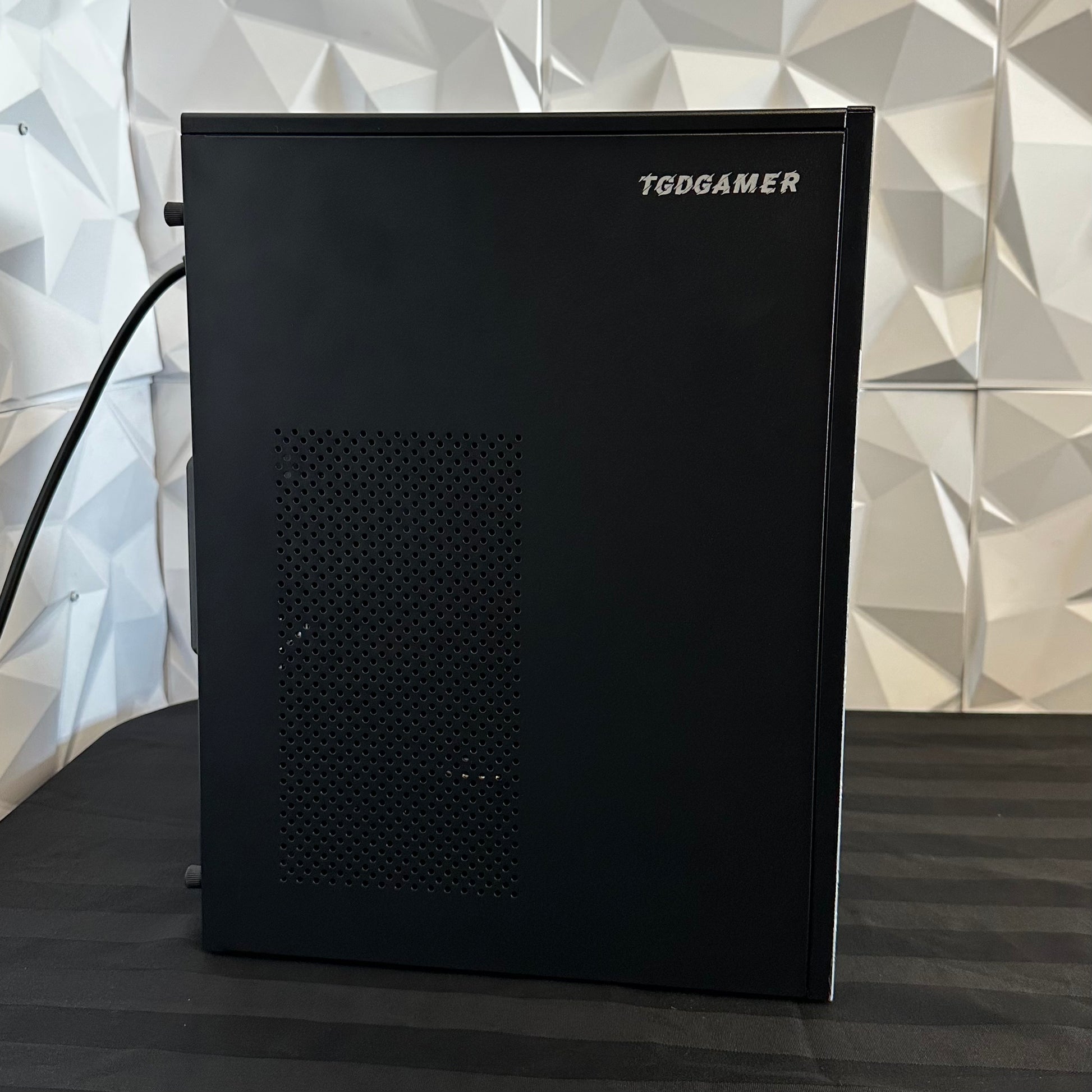 Black gaming computer tower with 'TGDGAMER' branding against a geometric patterned wall.