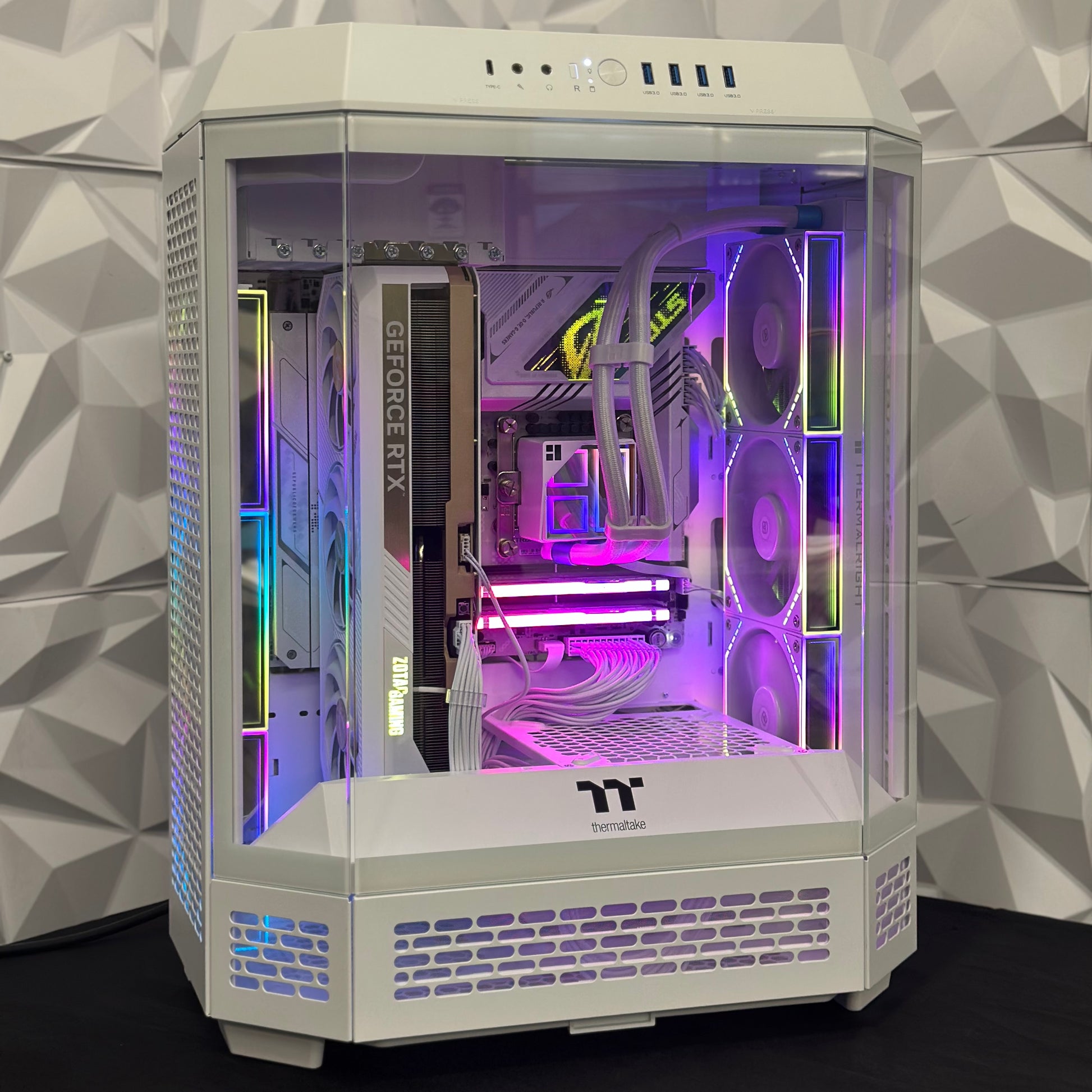 Computer gaming PC with RGB lighting inside a white case against a geometric-patterned wall.