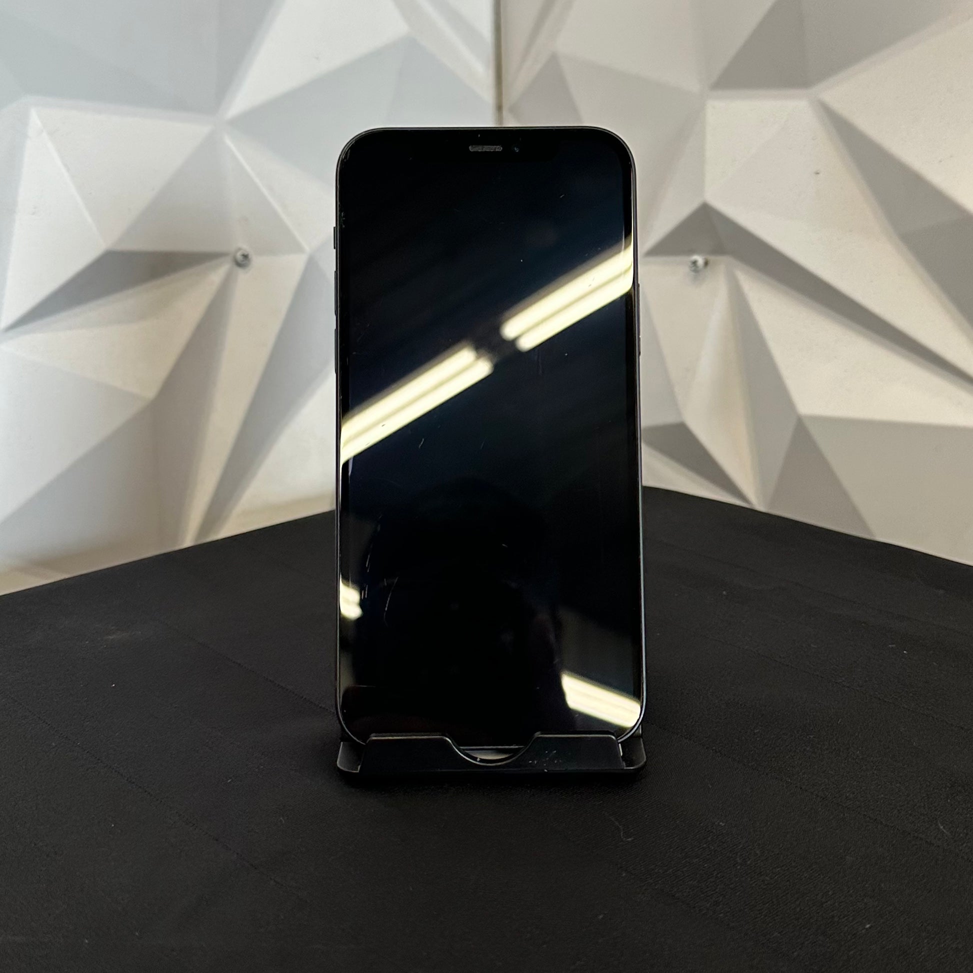 A black smartphone with a glossy screen standing upright on a black surface, set against a white geometric wall background.