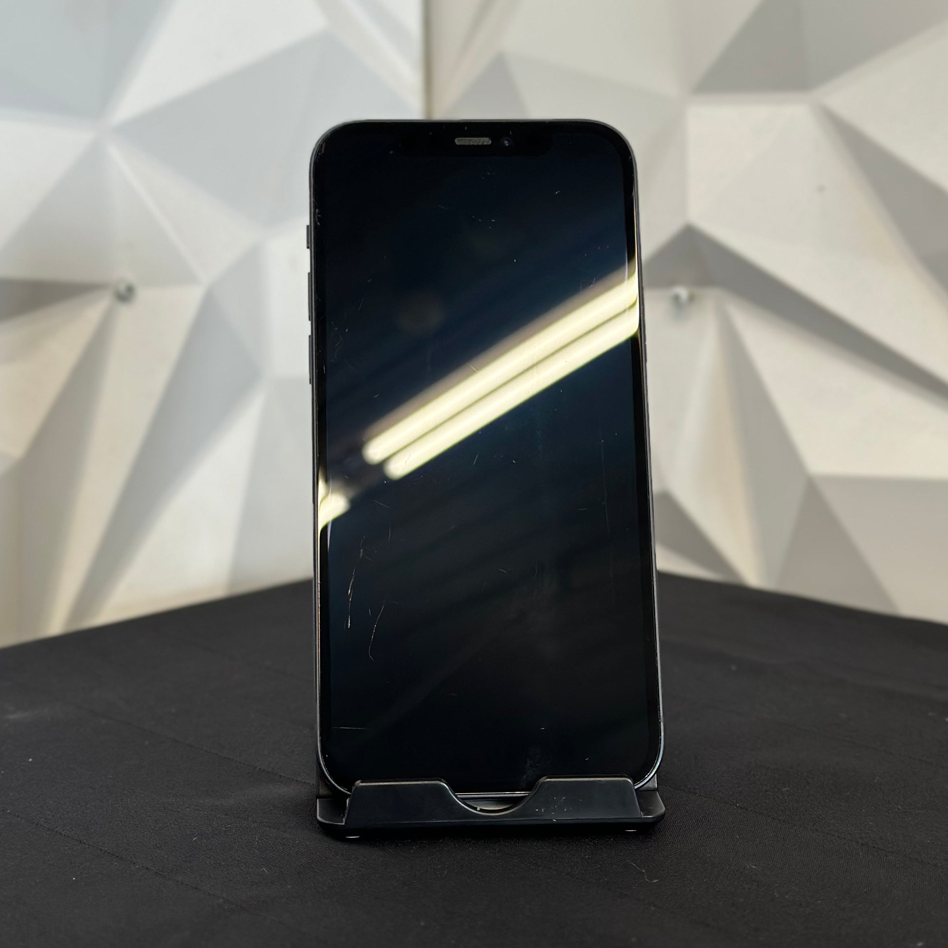 A black smartphone with a blank screen stands upright on a small display stand on a dark surface, with a geometric white wall background.