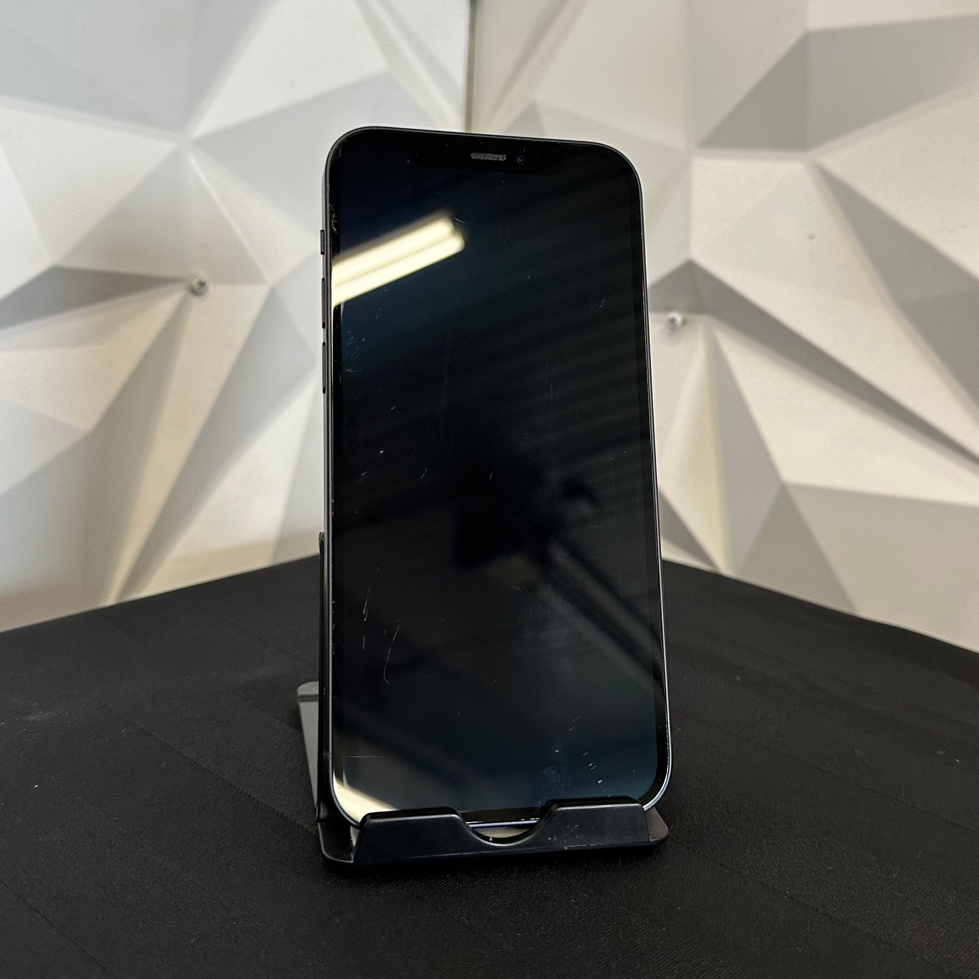 A black smartphone with a blank screen stands upright on a small black stand on a dark surface, with a white geometric wall behind it.