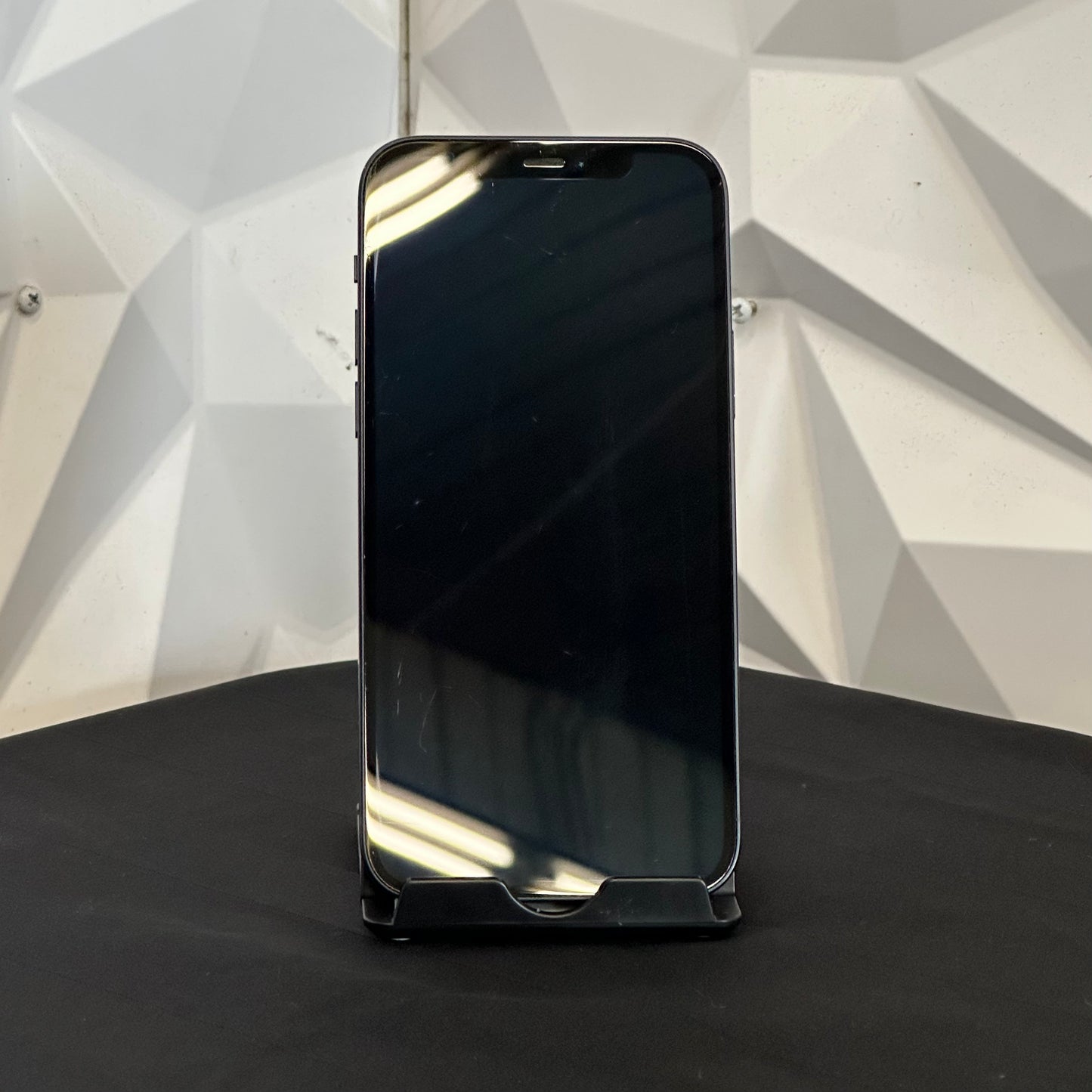 A black smartphone with a glossy screen, standing upright on a black surface against a white geometric textured wall.
