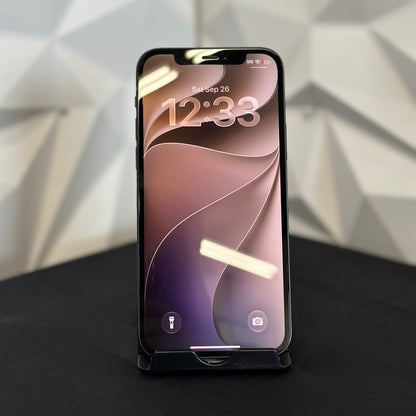 A black smartphone with a front-facing camera and a black protective bumper or case around the edges, standing upright on a black surface against a light gray geometric wall background.