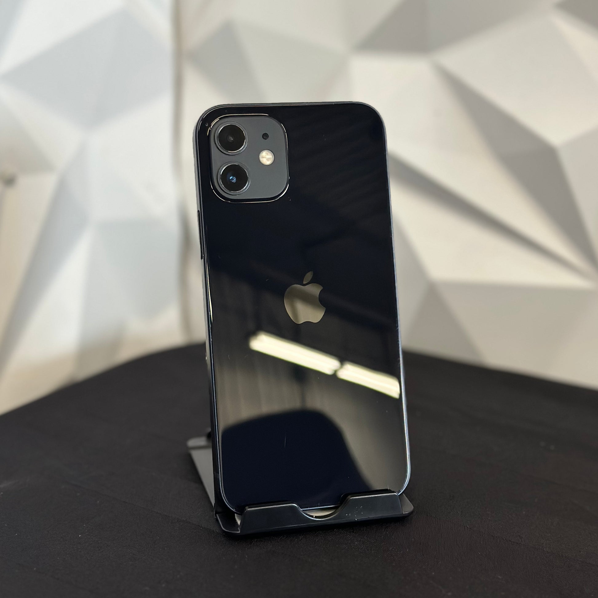 A black Apple iPhone 12 Pro shown from the back, resting on a black stand on a dark surface with a geometric white wall background.