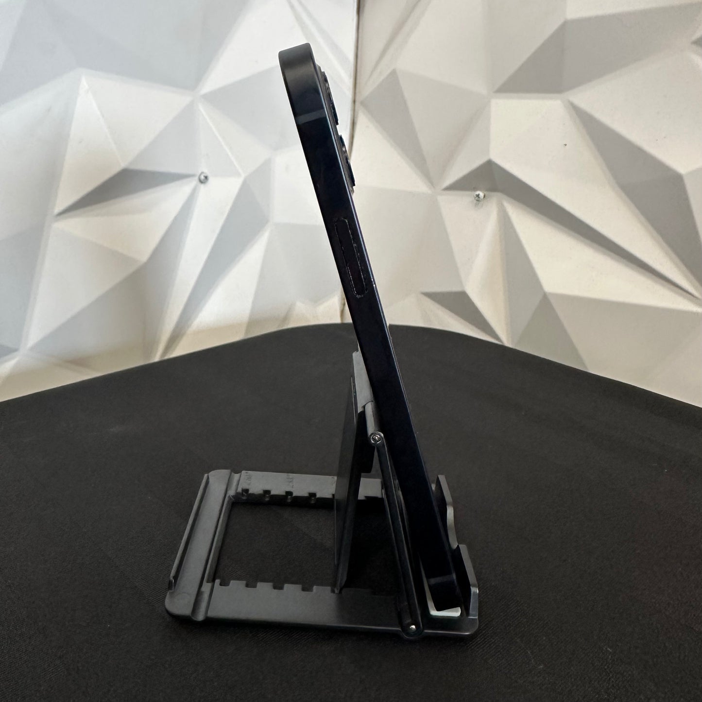 A black foldable smartphone stand holding a dark blue or black smartphone, set on a black surface against a white geometric faceted wall background.