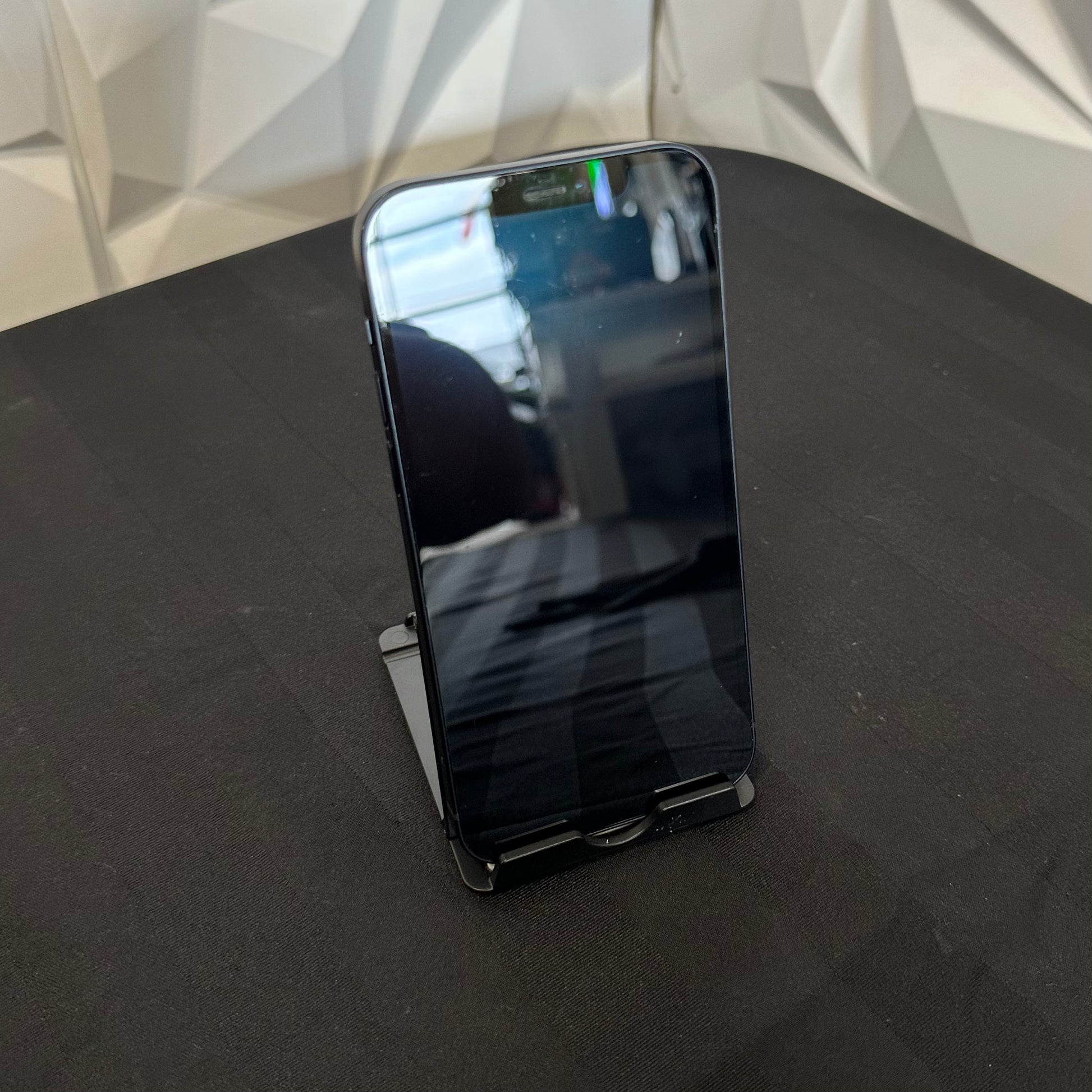 A black smartphone with a glossy screen stands upright on a small black stand on a dark tabletop, with a geometric white wall in the background.