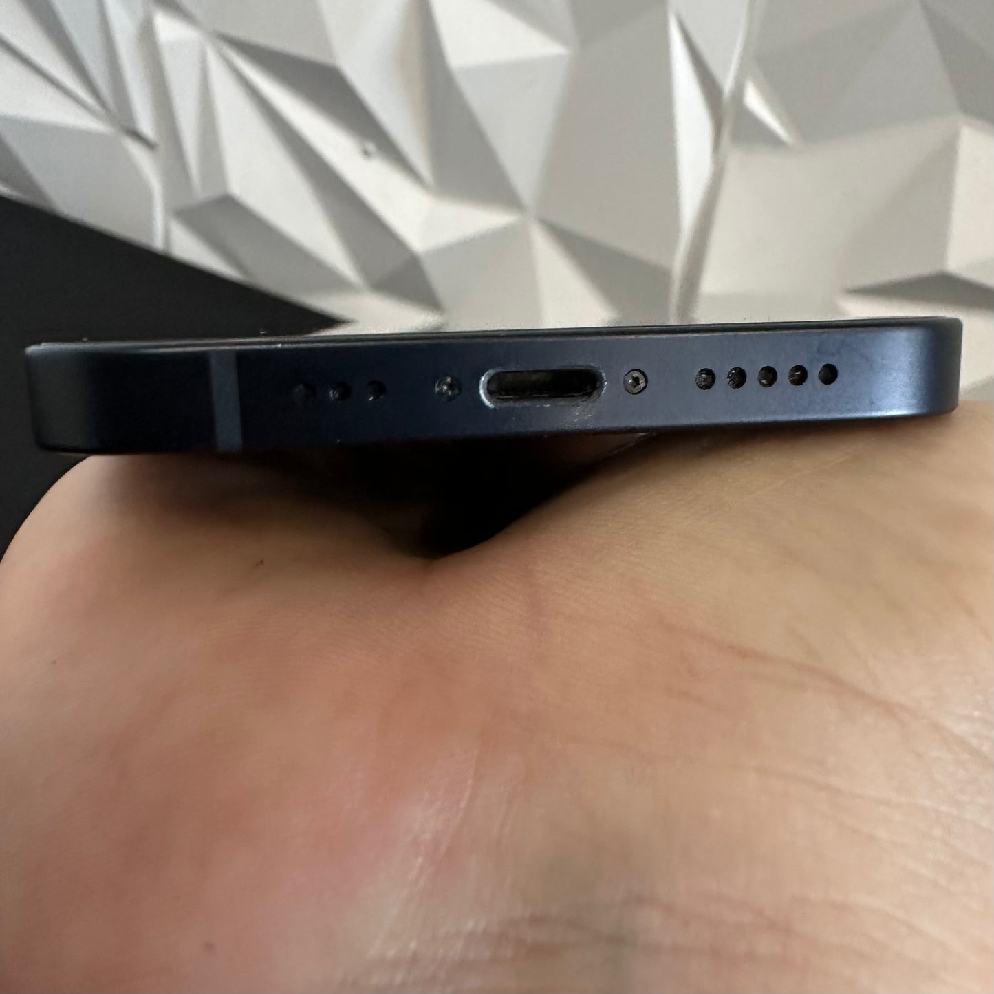 Close-up of the bottom edge of a dark blue smartphone being held in a hand, showing the USB-C charging port, speaker holes, and microphone cutouts against a geometric wall background.