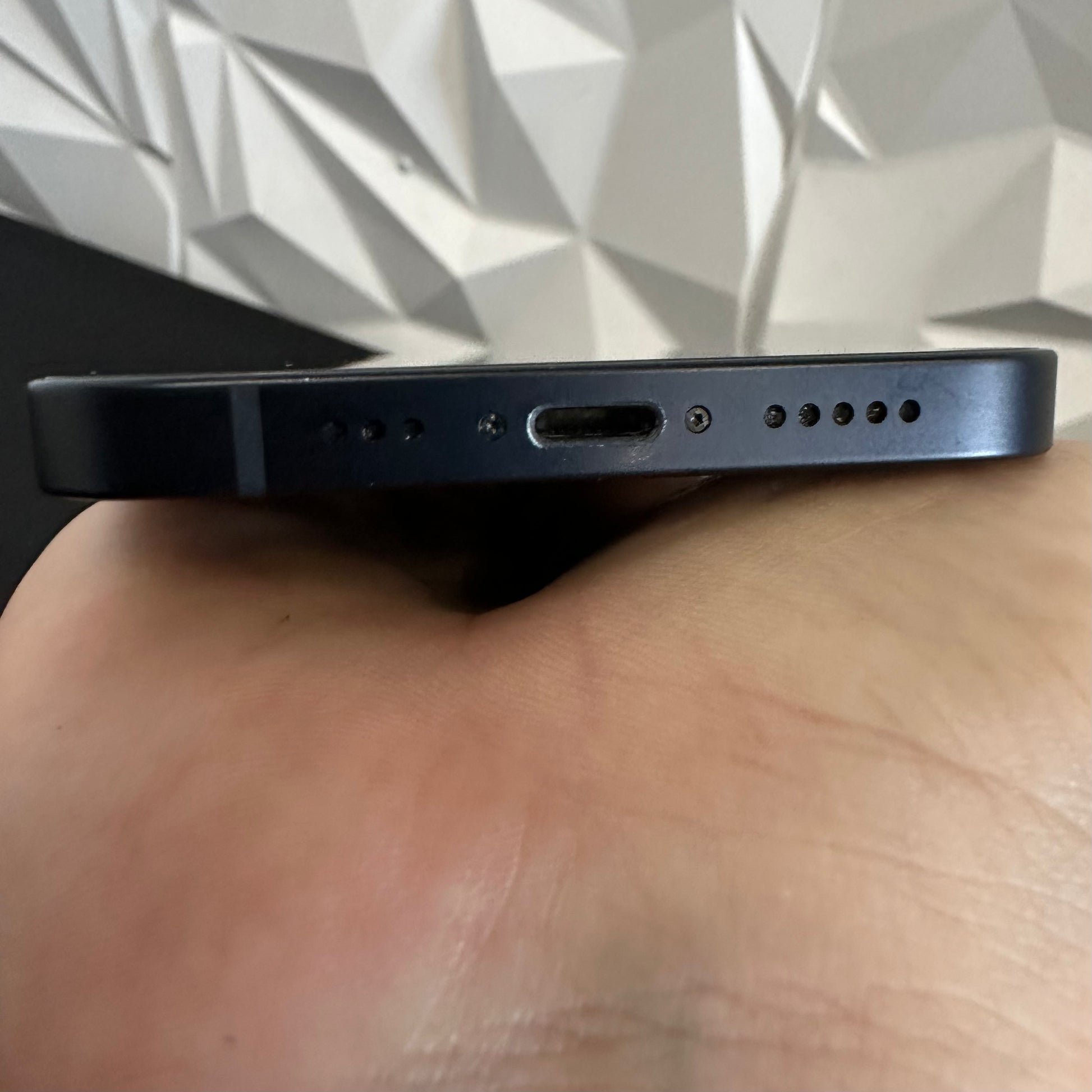 Close-up of the bottom edge of a dark blue smartphone being held in a hand, showing the USB-C charging port, speaker holes, and microphone cutouts against a geometric wall background.