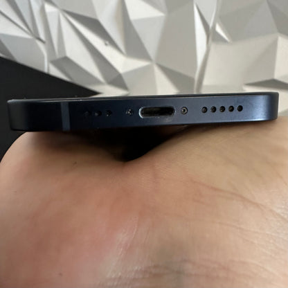 Close-up of the bottom edge of a dark blue smartphone being held in a hand, showing the USB-C charging port, speaker holes, and microphone cutouts against a geometric wall background.
